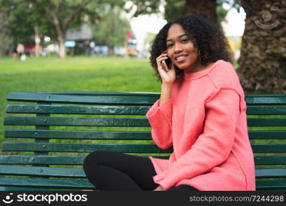 Portrait of young beautiful afro american woman talking on the phone and sitting on a bench in a park. Outdoors.