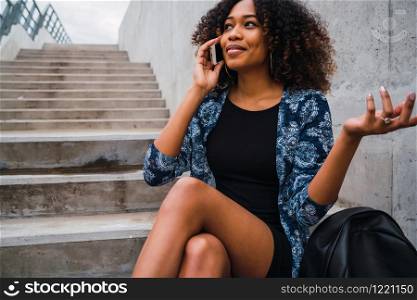 Portrait of young beautiful afro american woman talking on the phone while sitting on concrete steps outdoors.