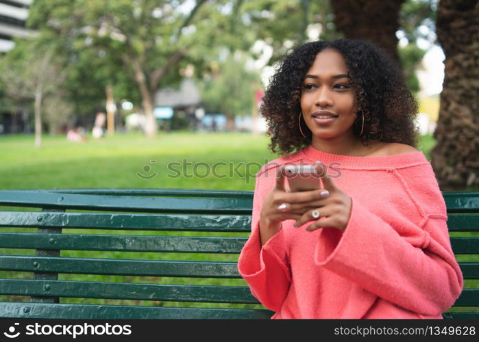 Portrait of young beautiful afro american woman using her mobile phone and sitting on a bench in a park. Outdoors.