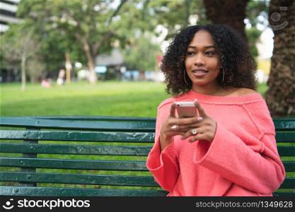 Portrait of young beautiful afro american woman using her mobile phone and sitting on a bench in a park. Outdoors.