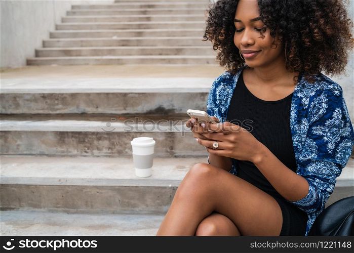 Portrait of young beautiful afro american woman using her mobile phone while sitting on concrete steps outdoors.