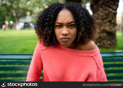 Portrait of young beautiful afro american woman with angry expression sitting on bench in the park. Outdoors.