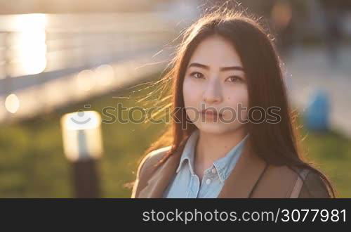 Portrait of young beautiful asian hipster woman with long brown straight hair looking at camera and smiling during sunset in backlight outdoors. Enigmatic smile of asian teenage female. Positive emotion and facial expression concept.