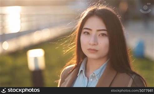 Portrait of young beautiful asian hipster woman with long brown straight hair looking at camera and smiling during sunset in backlight outdoors. Enigmatic smile of asian teenage female. Positive emotion and facial expression concept.