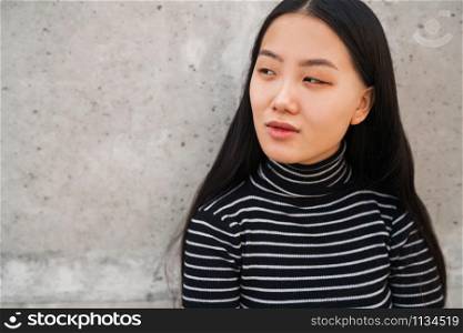 Portrait of young beautiful Asian woman standing outdoors against grey wall.