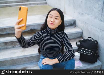 Portrait of young beautiful Asian woman taking a selfie with her mobile phone while sitting on concrete stairs outdoors.