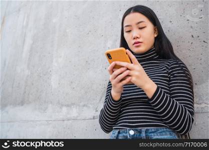 Portrait of young beautiful Asian woman using her mobile phone against grey wall. Communication concept.