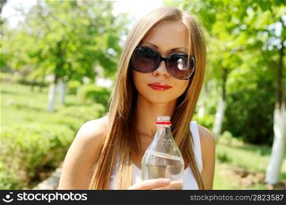 Portrait of young beautiful blond-haired woman wearing white t-shirt drinking water at summer green park.