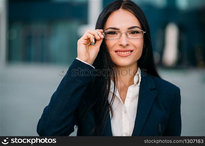 Portrait of young, beautiful business woman in glasses and suit. Modern building, financial center, cityscape. Successful female businessperson