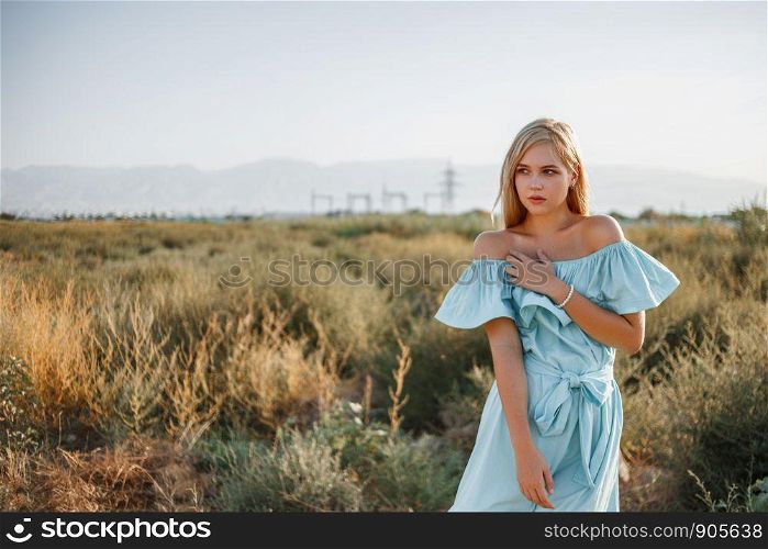 portrait of young beautiful caucasian blonde girl in light blue dress standing on the field with sun-dried grass during sunset