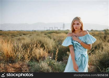 portrait of young beautiful caucasian blonde girl in light blue dress standing on the field with sun-dried grass during sunset