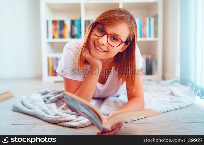 Portrait of young beautiful caucasian happy woman girl student reading or study at home lying on the floor in front of the book shelves wearing glasses holding a book smiling
