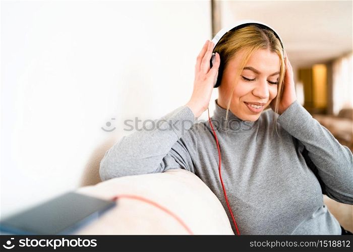 Portrait of young beautiful caucasian woman with earphones sitting in hotel - One alone adult happy blonde female listen to music in day - Waist up front view - Happiness enjoyment and joy concept