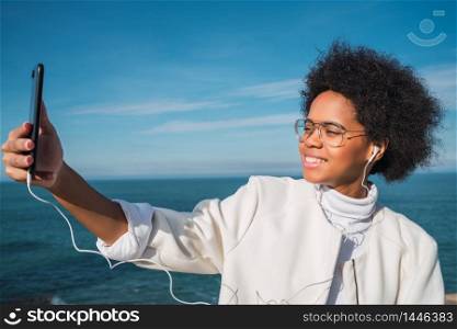 Portrait of young beautiful latin woman taking selfies with her mobile phone outdoors with the sea on background.