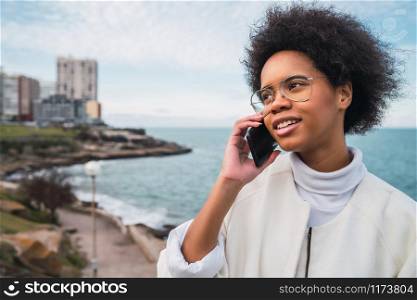 Portrait of young beautiful latin woman talking on the phone outdoors with the sea on background. Communication concept.
