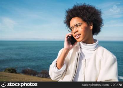 Portrait of young beautiful latin woman talking on the phone outdoors with the sea on background. Communication concept.