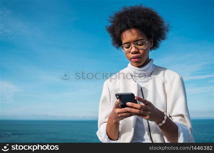 Portrait of young beautiful latin woman using her mobile phone outdoors with the sea on background. Technology and communication concept.