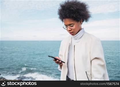 Portrait of young beautiful latin woman using her mobile phone outdoors with the sea on background. Technology and communication concept.