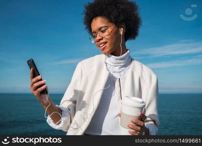 Portrait of young beautiful latin woman using her mobile phone with earphones and holding a cup of coffee, with the sea at background. Communication and technology concept.