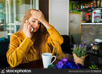 Portrait of young beautiful woman at cafe restaurant talking to the mobile phone call receiving disturbing surprising news holding her head with hand forehead