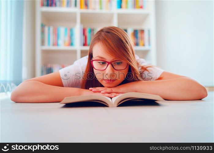 Portrait of young beautiful woman girl student reading or study at home lying on the floor in front of the book shelves wearing glasses holding a book