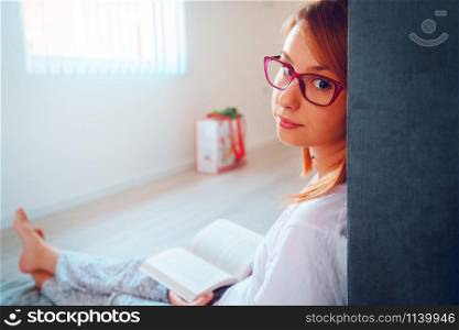 Portrait of young beautiful woman girl student reading or study at home lying on the floor in by the bed wearing glasses holding a book looking to the camera