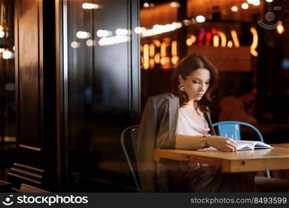 portrait of young beautiful woman is reading paper book in cafe in dusk. girl is online study, remote work. portrait of young beautiful woman is reading paper book in cafe in dusk. girl is online study, remote work.