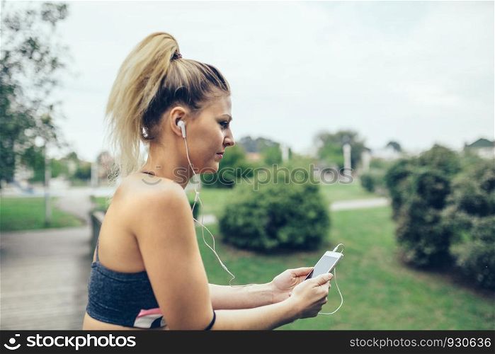 Portrait of young blonde woman with earphones listening music while looking her smartphone over nature background. Selective focus on phone.. Woman with earphones listening music in smartphone