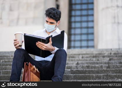Portrait of young business man wearing face mask and reading files while sitting on stairs outdoors. Business concept. New normal lifestyle concept.