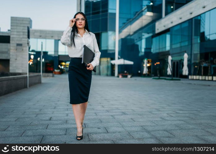 Portrait of young business woman in glasses and white blouse with notebook in hand, skyscaper on background. Modern building, financial center, cityscape. Successful female businessperson