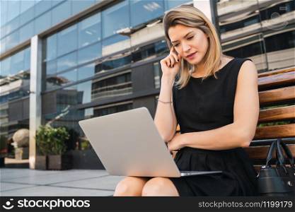 Portrait of young business woman using her laptop while sitting outdoors at the street. Business concept.