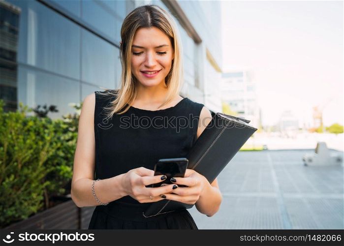 Portrait of young business woman using her mobile phone while standing outside office buildings. Business and success concept.