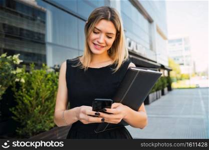 Portrait of young business woman using her mobile phone while standing outside office buildings. Business and success concept.
