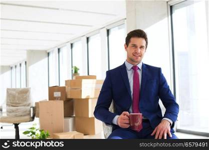 Portrait of young businessman having coffee with moving boxes in background at office