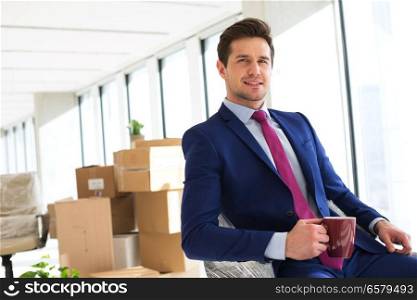 Portrait of young businessman holding coffee cup with moving boxes in background at office