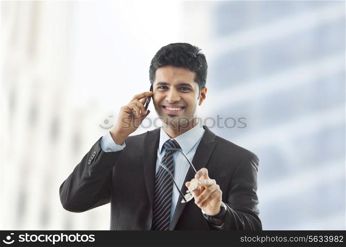 Portrait of young businessman holding glasses while using cell phone