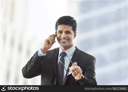 Portrait of young businessman holding glasses while using cell phone