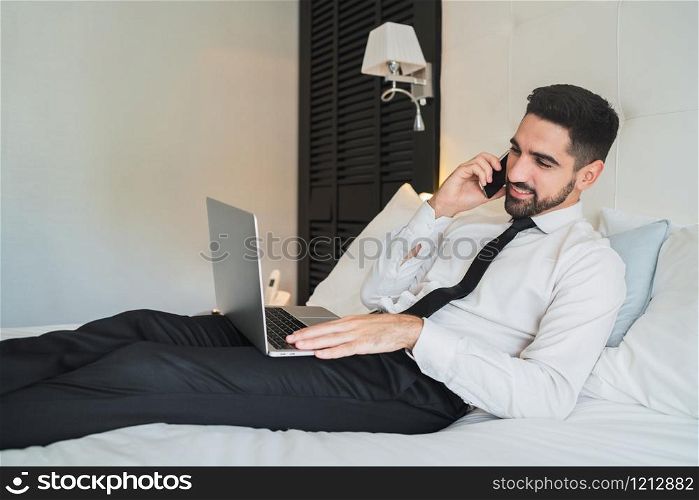 Portrait of young businessman lying on bed and talking on the phone while working on his laptop at the hotel room. Business travel concept.