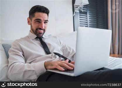 Portrait of young businessman lying on bed and working on his laptop at the hotel room. Business travel concept.