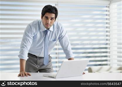 Portrait of young businessman standing by desk with laptop