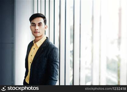 Portrait of Young Businessman Standing by the Window in Office. Looking at Camera