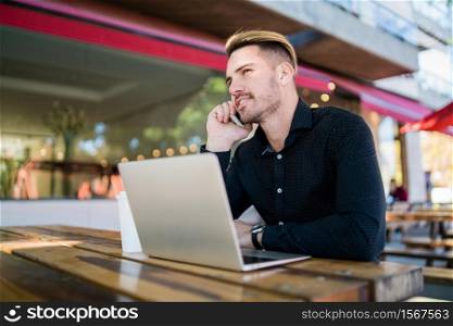 Portrait of young businessman working on his laptop and talking on the phone while sitting in a coffee shop. Technology and business concept.