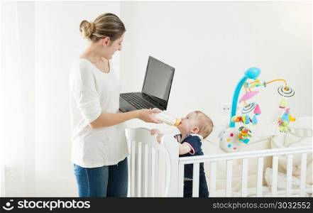 Portrait of young busy woman working on laptop and giving milk to her baby son. Portrait of busy woman working on laptop and giving milk to her baby son