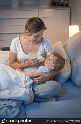 Portrait of young caring mother giving bottle with milk to her baby in bed at night. Parenting concept
