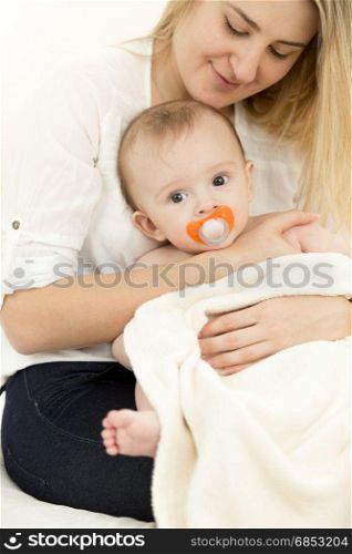 Portrait of young caring mother sitting on bed and holding baby on hands