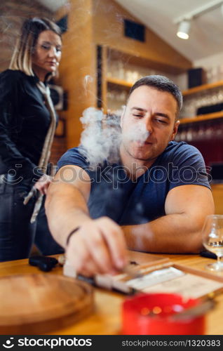 Portrait of young caucasian man adult sitting by table extinguishes a cigar smoking looking to ash tray wearing t-shirt blue in restaurant or home while unknown woman walk or stand behind him