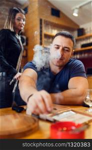 Portrait of young caucasian man adult sitting by table extinguishes a cigar smoking looking to ash tray wearing t-shirt blue in restaurant or home while unknown woman walk or stand behind him
