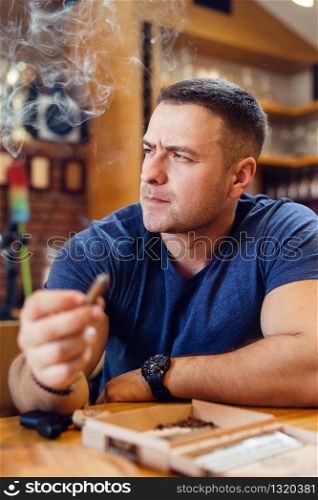 Portrait of young caucasian man adult sitting by the table holding a cigar smoking looking to the side wearing t-shirt blue in restaurant or home frowning thinking modern businessman