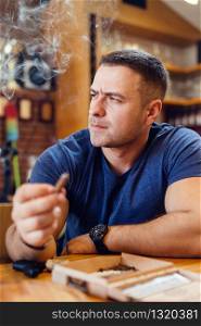 Portrait of young caucasian man adult sitting by the table holding a cigar smoking looking to the side wearing t-shirt blue in restaurant or home frowning thinking modern businessman