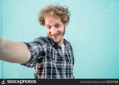 Portrait of young caucasian man looking at camera and taking a selfie against light blue background.